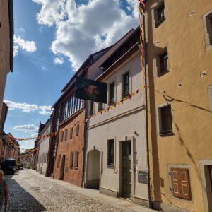 Gepflasterte Straße in der Altstadt Pirna mit bunten Wimpelketten und einem Bild der Künstlerin Yvonne Brückner sowie historischen Gebäuden.